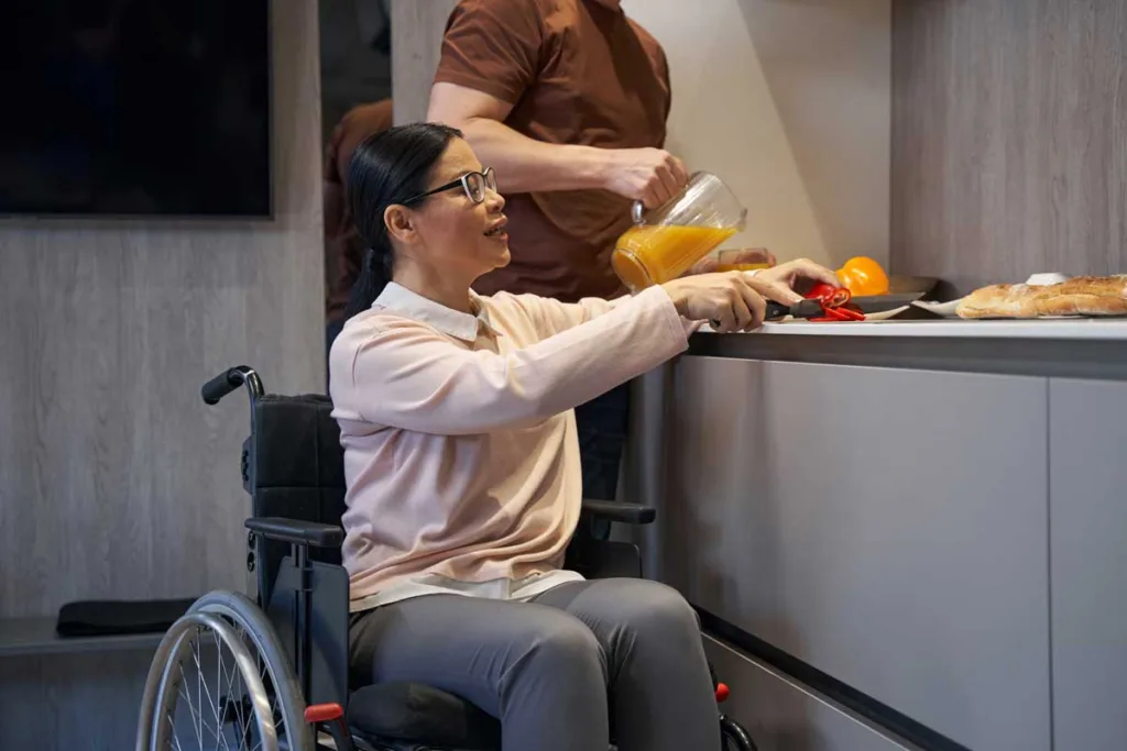 A woman in a wheelchair joyfully preparing breakfast in an accessible kitchen, reaching for a jug of orange juice.